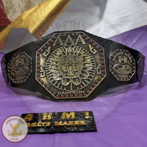 May include: A black leather wrestling championship belt with gold and red accents. The belt features the letters "AAA" and the word "CAMPEON". The center plate has a detailed gold design. The belt is on a purple surface.