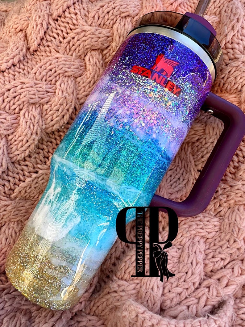 K&ouml;nnte beinhalten: Ein individueller Stanley-Becher mit lila Griff und Strohhalm. Der Becher ist mit einem glitzernden Ozean-Design in Blau-, T&uuml;rkis-, Lila- und Goldt&ouml;nen verziert. Das Stanley-Logo ist rot. Das Preppy Pepper-Logo ist ebenfalls sichtbar.