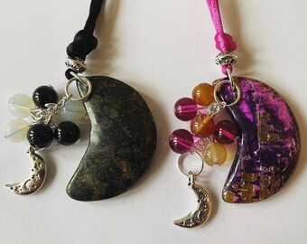 May include: Two crescent moon shaped pendants with silver chains and charms. One pendant is green with black and white beads, the other is purple with orange and red beads.