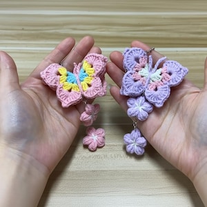 Pode incluir: Dois brincos de borboleta de crochê. Um é rosa com detalhes amarelos e azuis, o outro é roxo com detalhes rosa e branco. Cada brinco tem pingentes de flores pendurados. Os brincos são feitos à mão.