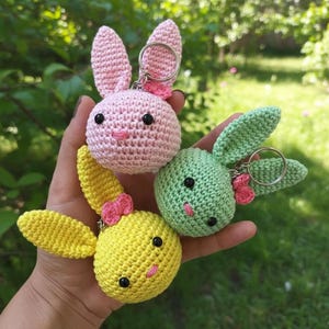 Puede incluir: Tres llaveros de conejito hechos a mano en crochet, rosa, verde y amarillo. Cada conejito tiene orejas largas, ojos negros y un lazo rosa. Los llaveros son redondos y tienen un anillo metálico para sujetar. Fondo de follaje verde borroso.