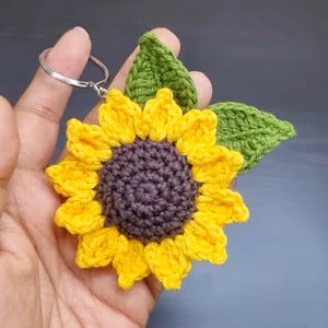 Puede incluir: Un llavero de girasol tejido a ganchillo. El girasol tiene pétalos amarillos, un centro marrón y hojas verdes. El llavero se sostiene en una mano, mostrando los intrincados detalles del trabajo con hilo. Un accesorio encantador, inspirado en la naturaleza.
