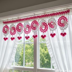 May include: Crocheted window valance in white and pink. The valance features a scalloped top edge and decorative circular motifs with dangling pink pom-poms. The design is displayed against a backdrop of white curtains and a window.