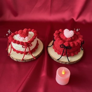 May include: Two heart-shaped crochet cakes, one white and red, the other all red, sit on wooden slices. The cakes are decorated with hearts and bows. A pink candle with a flickering yellow light is in the foreground. The background is a red satin fabric.
