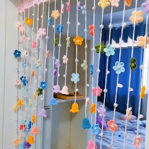 May include: A colourful, handmade crochet door curtain. It features a variety of crocheted flowers and bell shapes in shades of pink, blue, yellow, and orange. The curtain is made of white string with decorative pearls interspersed along the strings.