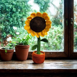 May include: A handcrafted crochet sunflower in an orange pot sits on a wooden windowsill. The sunflower has a brown center and yellow petals. Green leaves and stem complete the design. Other potted plants are also visible.