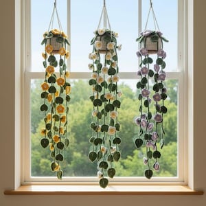 Może przedstawiać: Trzy wiszące donice z szydełkowymi pnączami i kwiatami. Donice zawieszone są przed oknem. Jedna donica ma żółte kwiaty, druga białe, a trzecia fioletowe. Pnącza mają zielone liście.