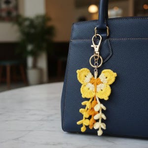 May include: A yellow and white crochet butterfly bag charm with a gold-colored clasp is attached to a navy blue handbag. The butterfly has a detailed design, and the charm includes a cascade of crochet flowers in shades of yellow and cream.