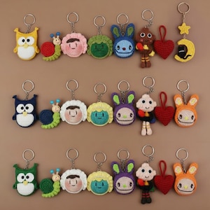 May include: A collection of colorful, handmade crochet keychains. The keychains feature various designs, including an owl, snail, faces, a bunny, a doll, a heart, and a moon and star. The keychains are made with yarn and metal key rings.
