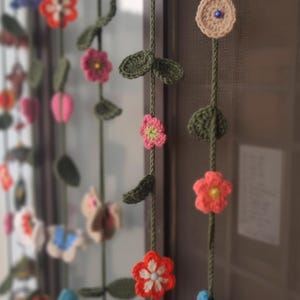 Puede incluir: Guirnalda floral de ganchillo hecha a mano con varias flores coloridas y hojas verdes. La guirnalda está ensartada en un cordón verde, con flores en tonos de rojo, rosa y beige. Un artículo decorativo encantador, hecho a mano.