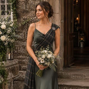 Puede incluir: Un vestido largo de satén verde oliva con escote drapeado y tirantes finos, que presenta una banda de tartán a cuadros oscuros sobre un hombro, sujeta con un broche circular plateado, acompañado de un ramo de flores blancas.