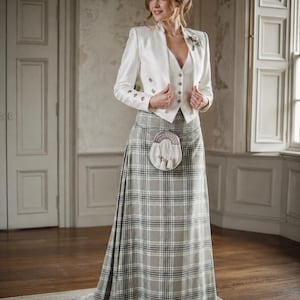 Puede incluir: Una chaqueta blanca entallada con botones plateados sobre un chaleco blanco a juego, combinada con una falda kilt larga de cuadros grises y blancos. Un esporrán de cuero blanco con herrajes plateados cuelga de un cinturón de cadena.