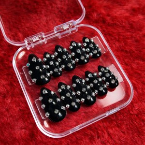 Peut inclure: Un ensemble de faux ongles noirs en forme d'amande, chacun orné de plusieurs petits strass étincelants. Les ongles sont présentés dans un étui en plastique transparent sur un fond rouge texturé.
