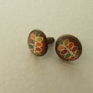 Peut inclure: Une paire de boucles d'oreilles rondes avec un motif botanique. Les boucles d'oreilles présentent une base marron foncé avec un motif de feuilles colorées en orange, vert, jaune et blanc. Le sertissage métallique est de couleur bronze.