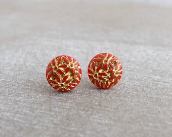 Può includere: Un paio di orecchini a lobo rotondi caratterizzati da una finitura in smalto rosso brillante con un motivo floreale in rilievo color oro metallizzato. Il design circolare mostra molteplici forme di fiori sulla superficie, creando un aspetto strutturato.