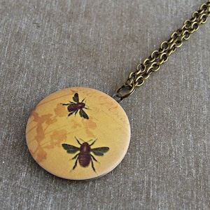 May include: A circular pendant necklace with a yellow backdrop, showcasing two illustrated bees. The pendant is suspended from a bronze-toned chain. The bees are brown and black.