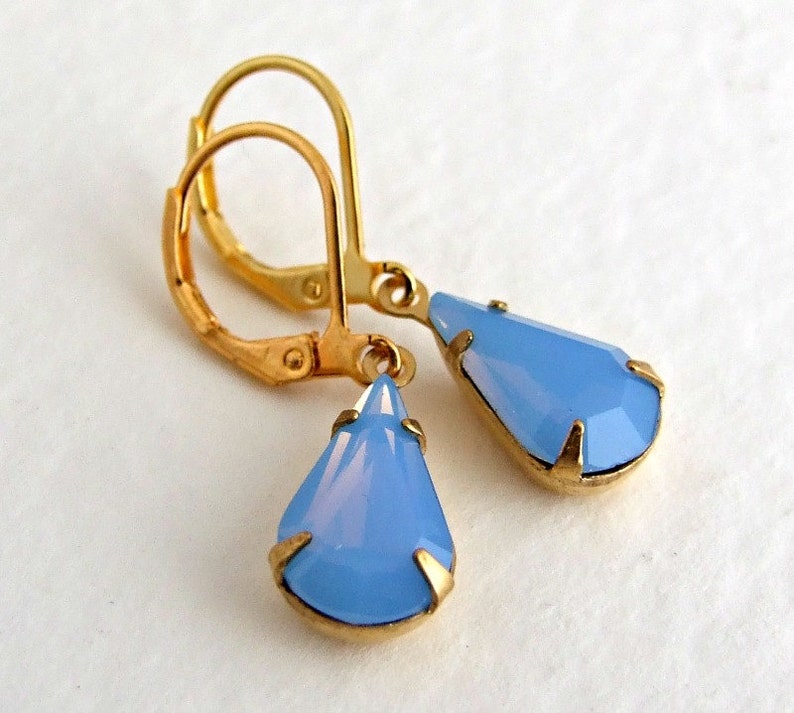 May include: A pair of gold-colored leverback earrings with teardrop-shaped, light blue pendants. The pendants are faceted and set in gold-colored metal. The earrings are displayed against a white background.