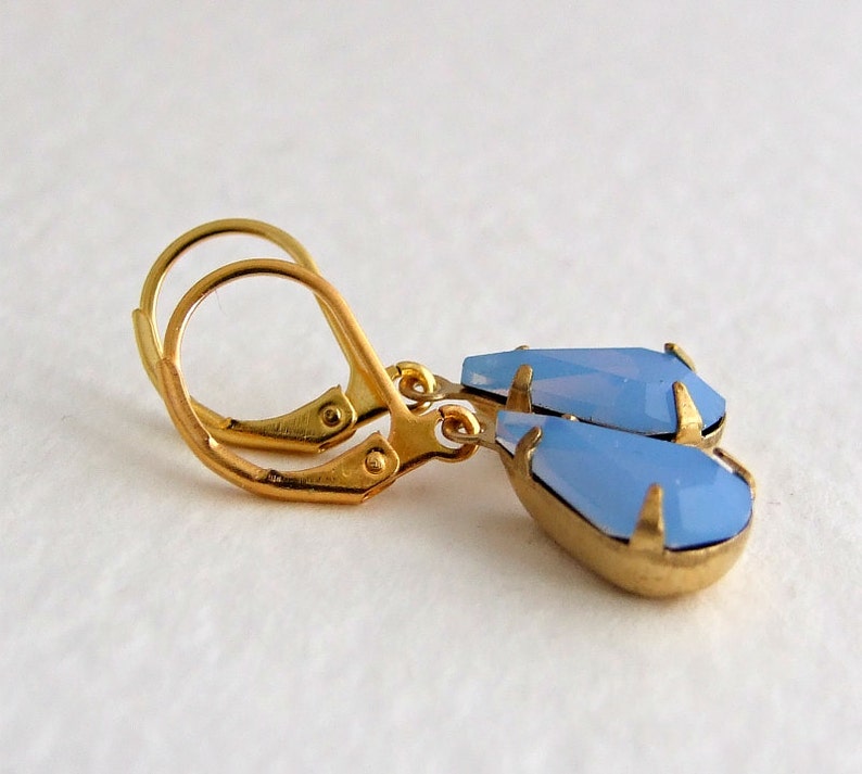May include: A pair of gold-toned leverback earrings with teardrop-shaped, light blue gemstones. The earrings have a faceted design, and the gemstones are set in a gold-colored metal. The earrings are displayed against a white background.