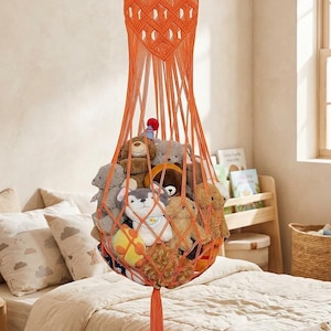 May include: An orange macrame hanging storage basket filled with stuffed animals. The basket is suspended from a wooden dowel and features a diamond pattern at the top. A tassel hangs from the bottom. The basket is filled with various plush toys.