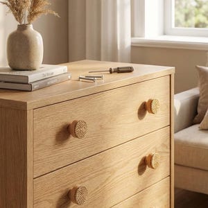 May include: A light oak dresser with four drawers, each featuring a woven knob. A vase with dried plants sits atop the dresser, along with books, screws, and a screwdriver. The room is lit by natural light.