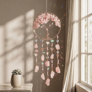 May include: A rose gold-colored dream catcher with a tree of life design. The circular frame is adorned with pink crystals and a wire tree. Strands of pink crystals, butterflies, and raw rose quartz hang below. A succulent in a white pot sits on a nearby table.