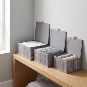 May include: Three gray storage boxes with hinged lids, made of a bamboo-like material, sit on a wooden surface. The boxes are open, revealing folded white and gray towels. The boxes are of varying sizes, offering versatile storage solutions.