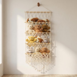 May include: A macrame wall hanging with multiple wooden shelves, displaying a collection of plush stuffed animals. The animals include teddy bears, dogs, and bunnies in various colors. The wall hanging is suspended from a wooden peg.
