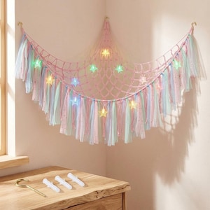 May include: A pink macrame net with colorful ribbons and star-shaped lights. The net is hung on a wall and decorated with pastel-colored ribbons in shades of pink, blue, and green. The star lights are in various colors.