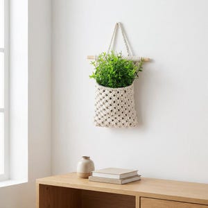 May include: A cream-colored macrame plant holder with a wooden dowel and hanging cord, filled with vibrant green foliage. The woven design adds a touch of bohemian style. The plant holder is suspended against a white wall.