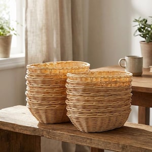 May include: Two woven wicker baskets, one stacked on top of the other, sit on a rustic wooden bench. The baskets are a light tan color and have a rounded shape. The background includes a window, curtains, and a wooden table.