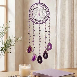 May include: A purple dreamcatcher with a circular frame adorned with small purple stones and hanging strands of purple beads and agate slices. A lit candle, a lavender sprig, and a purple journal are on a wooden surface.