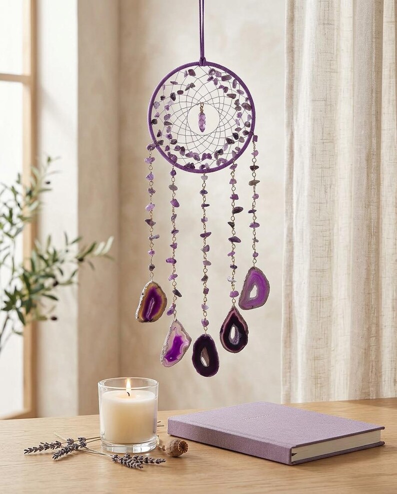 May include: A purple dreamcatcher with a circular frame adorned with small purple stones and hanging strands of purple beads and agate slices. A lit candle, a lavender sprig, and a purple journal are on a wooden surface.