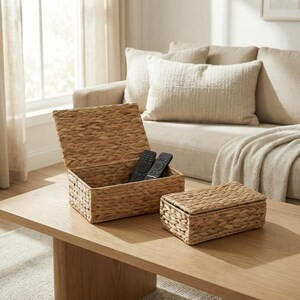 May include: Two woven water hyacinth storage boxes, one open and one closed, sit on a wooden coffee table. The open box contains two black remote controls. The boxes are a natural tan color and have a rectangular shape.