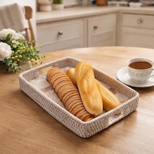 May include: A rectangular woven basket with handles holds a variety of baked goods, including sliced bread and baguettes. The basket sits on a wooden table, next to a cup of coffee and a floral arrangement. The basket is beige.