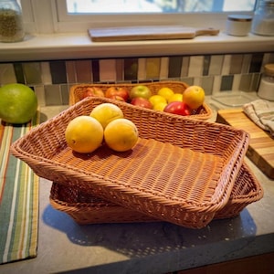 May include: Two rectangular woven baskets with a light brown finish, stacked one inside the other. The top basket contains three yellow citrus fruits. The baskets feature a consistent, tight weave pattern throughout the base and sides.
