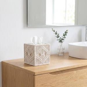 May include: A square tissue box cover made of woven beige macrame. The cover features a geometric diamond pattern and holds white tissues. The box sits on a light wood surface in a bathroom setting.