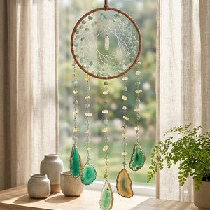 May include: A handcrafted dreamcatcher with a brown wooden hoop, adorned with green and yellow gemstones. The dreamcatcher features hanging strands of gemstones and large, polished agate slices in shades of green and gold. It hangs in front of a window.