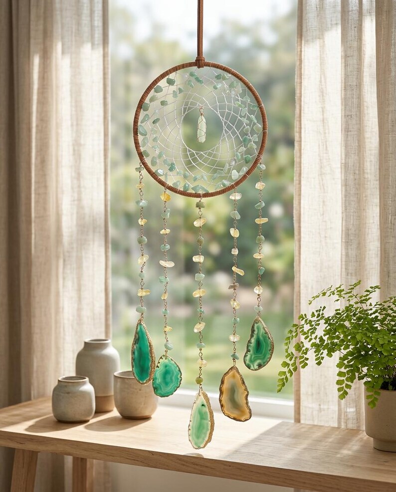 May include: A handcrafted dreamcatcher with a brown wooden hoop, adorned with green and yellow gemstones. The dreamcatcher features hanging strands of gemstones and large, polished agate slices in shades of green and gold. It hangs in front of a window.