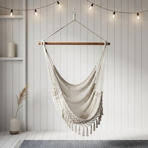 May include: A cream-colored macrame hammock chair with a wooden bar and fringe detail. The chair hangs from a metal ring and rope, against a white slatted wall. String lights are strung above the chair.