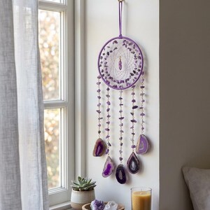 May include: A purple dreamcatcher with amethyst and agate accents hangs on a wall. The circular frame is adorned with small amethyst stones and dangling strands of amethyst beads and agate slices. A gold hook suspends the dreamcatcher near a window.