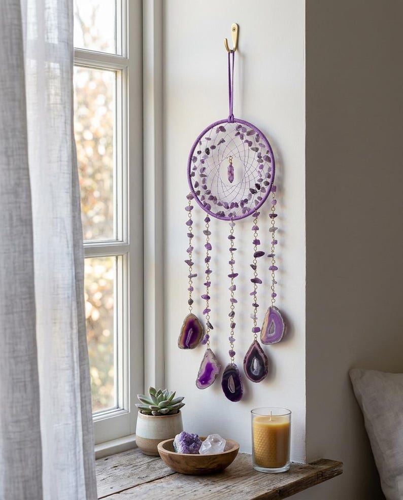 May include: A purple dreamcatcher with amethyst and agate accents hangs on a wall. The circular frame is adorned with small amethyst stones and dangling strands of amethyst beads and agate slices. A gold hook suspends the dreamcatcher near a window.