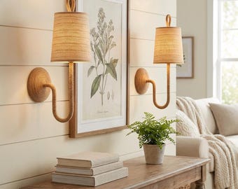 May include: Two wall sconces with beige fabric shades and rope-wrapped arms and backplates. The sconces are mounted on a light-colored wall. A framed botanical print hangs between the sconces. A wooden table holds books and a potted plant.