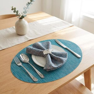 May include: A teal oval placemat holds a white plate with a folded blue and tan patterned cloth napkin secured by a silver ginkgo leaf napkin ring. A silver fork, spoon, and knife are arranged on the placemat beside the plate.