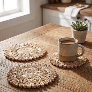 May include: Three woven, circular coasters with a scalloped edge sit on a wooden table. A beige ceramic mug filled with a brown liquid rests on one of the coasters. A small potted succulent is in the background.