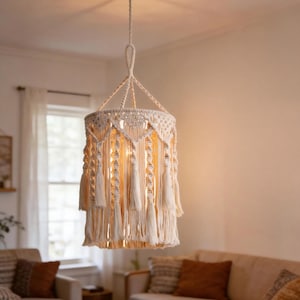 May include: Cream-colored macrame pendant light fixture with a cylindrical shape. The light has a woven design with diamond patterns and long tassels hanging from the bottom. The fixture is suspended from the ceiling, illuminating a living room.