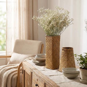 May include: A rectangular woven vase filled with white flowers sits on a wooden table. A smaller, conical woven vase and two bowls are also on the table. A chair and a blanket are in the background.