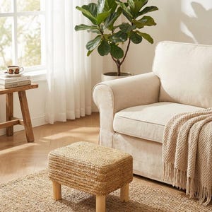 May include: A woven, rectangular ottoman with light wood legs sits on a jute rug. A beige sofa with a fringed throw blanket and a potted plant are in the background. The room has a neutral color palette.