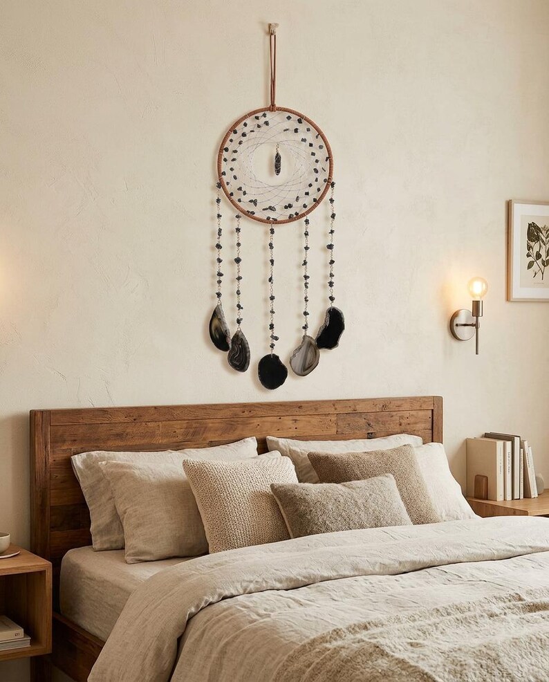 May include: A brown and black dreamcatcher with black stone beads and agate slices hanging below a circular frame. The dreamcatcher is suspended above a bed with neutral-toned bedding and pillows.