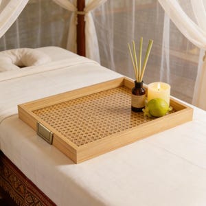May include: A rectangular wooden tray with a woven cane base and metallic side handles. The tray holds a small amber glass bottle with reed diffusers, a lit white pillar candle, and a single green lime on a white massage table.