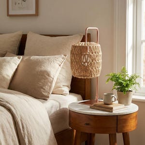 May include: A bedroom scene featuring a woven lamp with a copper frame on a wooden nightstand with a marble top. The bed has beige linens and pillows. A small potted plant and a mug are on the nightstand.